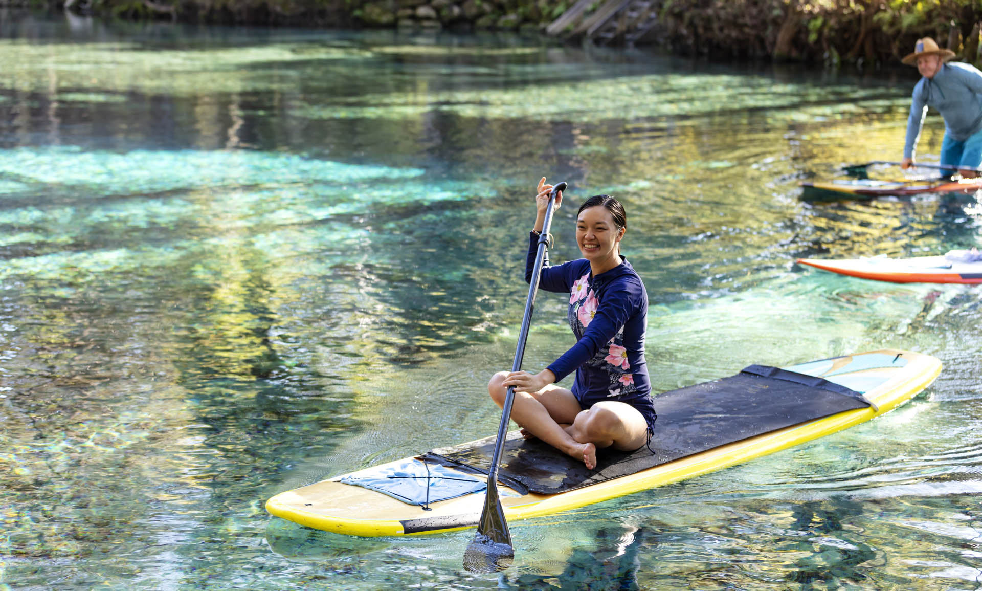 A woman sitting crossed-legged on a stand up paddleboard. She is paddleboarding on a tranquil Florida river enjoying nature. She is smiling and looking at the camera.