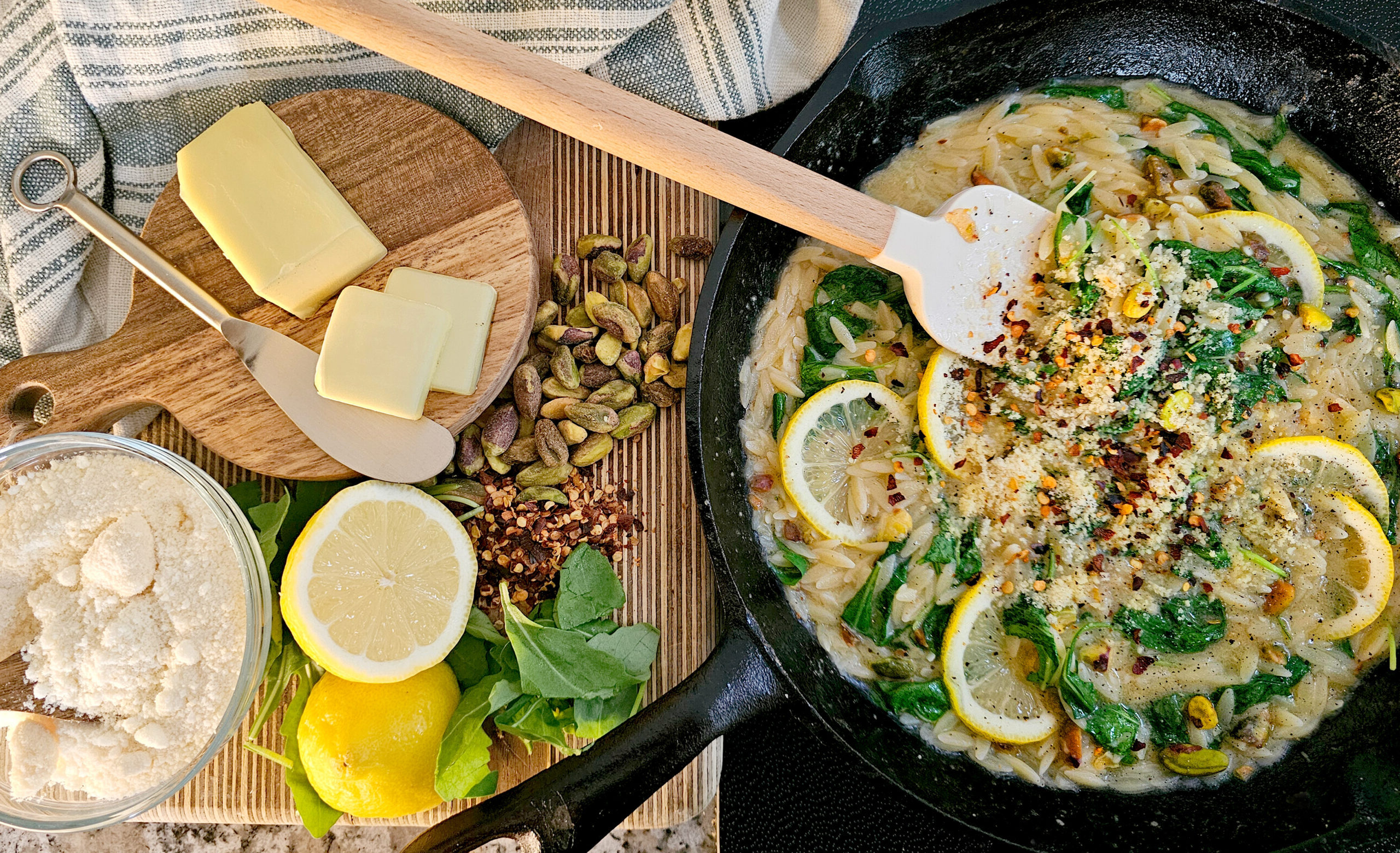 A cast-iron skillet filled with creamy lemon orzo pasta, garnished with spinach, lemon slices, pistachios, red pepper flakes, and parmesan. A spatula rests in the skillet. To the side, a wooden board holds butter slices, whole pistachios, red pepper flakes, fresh lemons, spinach leaves, and a bowl of grated parmesan cheese. A striped kitchen towel is draped nearby.