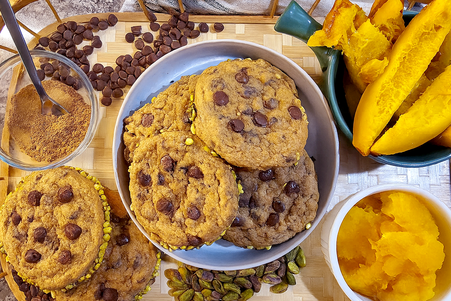 GRO-Pumpkin Cookies 3 freshly baked pumpkin chocolate chip cookies with crushed pistachios around the rims, displayed on a bamboo tray. The scene includes roasted pie pumpkin slices, a bowl of pumpkin puree, chocolate chips, pistachios, and a small dish of pumpkin pie spice, all captured in warm natural light on a granite countertop.