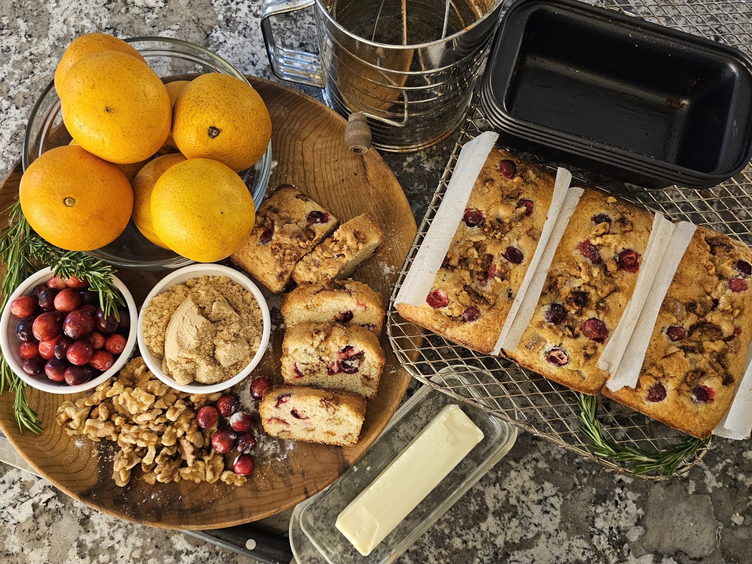 Overhead view of freshly baked cranberry orange walnut quick bread, sliced and cooling on a wire rack. Whole oranges, fresh cranberries, brown sugar, walnuts, rosemary sprigs, and butter are arranged on a wooden board beside the loaf on a stone countertop.