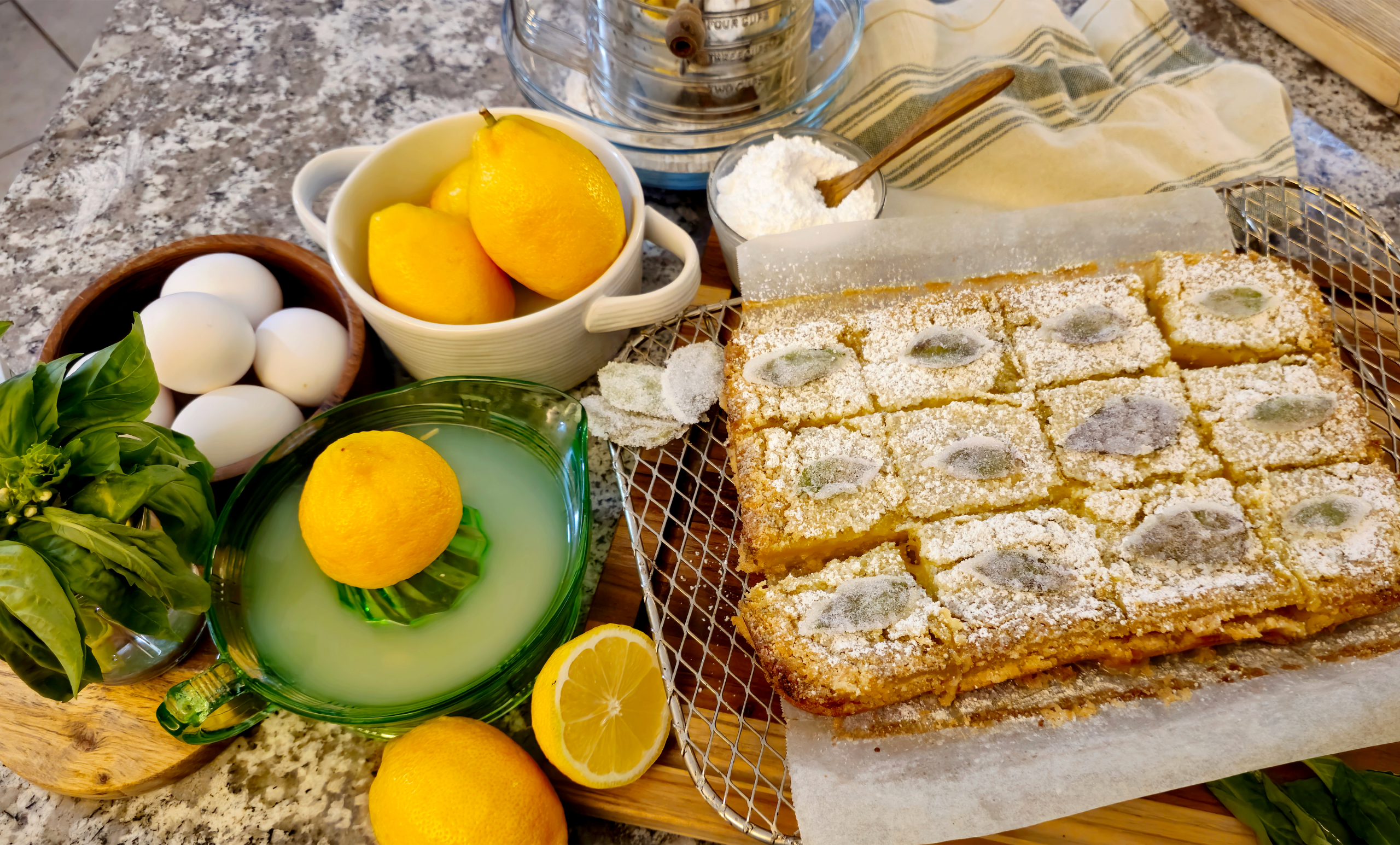 An overhead view of fresh baked lemon basil bars, cut into squares and cooling on a wire rack atop a wooden board. Whole and halved lemons, eggs, confectioner's sugar, basil leaves, and a juicer with lemon juice are artfully arranged next to the bars, on a stone countertop.