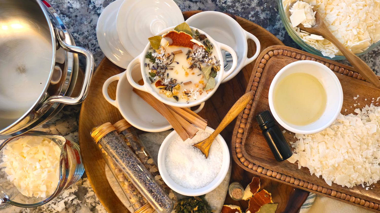 An overhead view of homemade soy candles. The setup shows the making process on a kitchen countertop. A small white ceramic vessel filled with melted soy wax is topped with dried citrus slices, herbs, and botanicals, surrounded by bowls of soy wax flakes, essential oil, coarse salt, cinnamon sticks, and dried botanicals. A stainless steel pot, wooden trays, measuring bowls, and glass jars are neatly arranged, creating a warm, rustic scene.