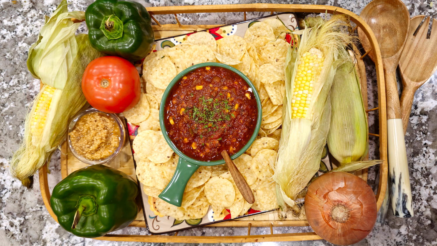 An overhead view of a bowl of simmered garden salsa, surrounded by nacho chips and ears of corn, arranged on a ceramic plate and wooden tray that sit atop a granite counter. Ingredients like green bell peppers, tomatoes, an onion, and a ripe tomato, and are artfully arranged around the chips. A wooden serving spoon and fork sit to the right.