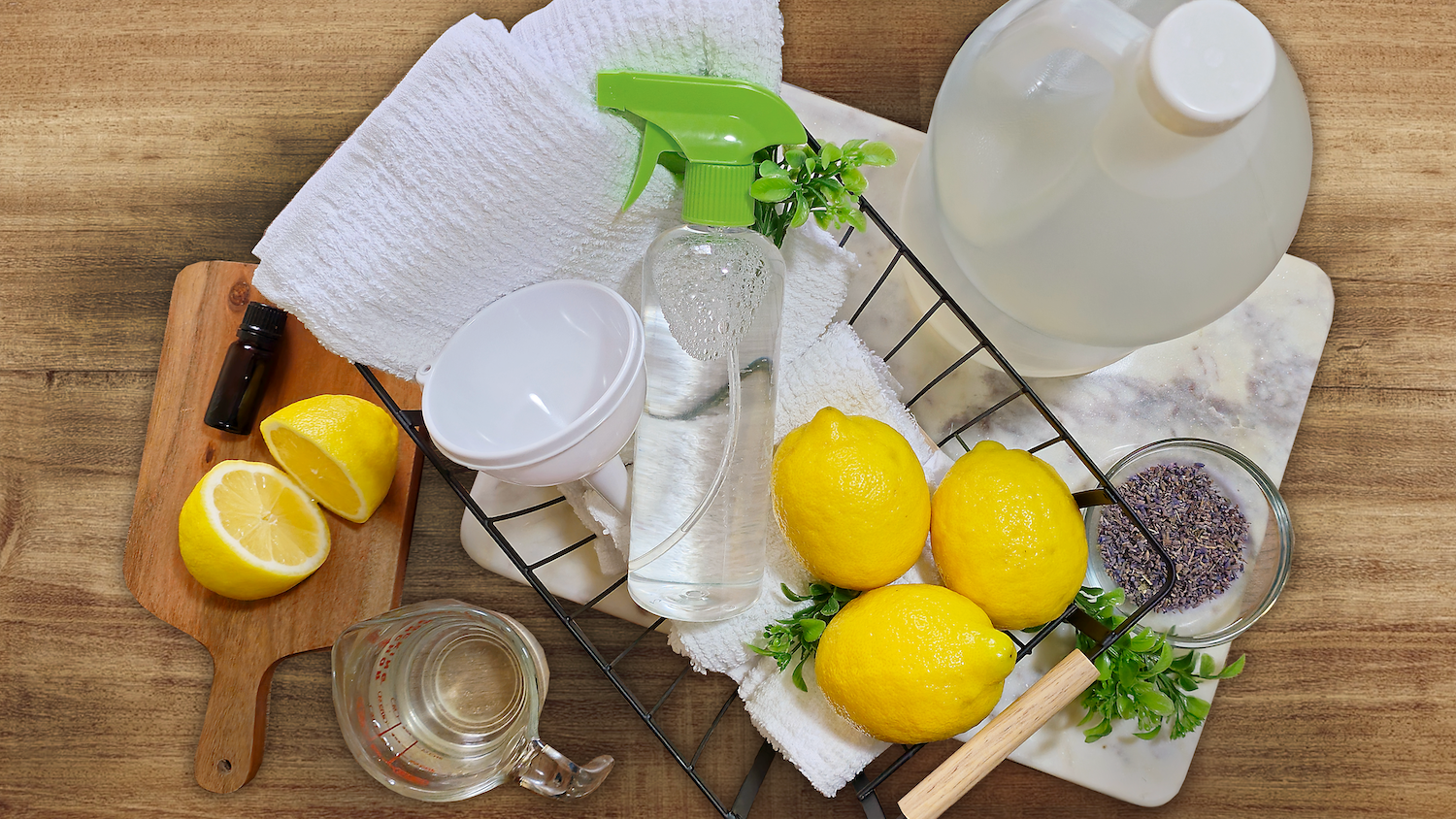 Overhead view of DIY all-natural cleaner supplies including lemons, a spray bottle with liquid, a jug of vinegar, a measuring cup, a small bottle of essential oil, dried lavender, a funnel, and white towels arranged on a wooden surface.