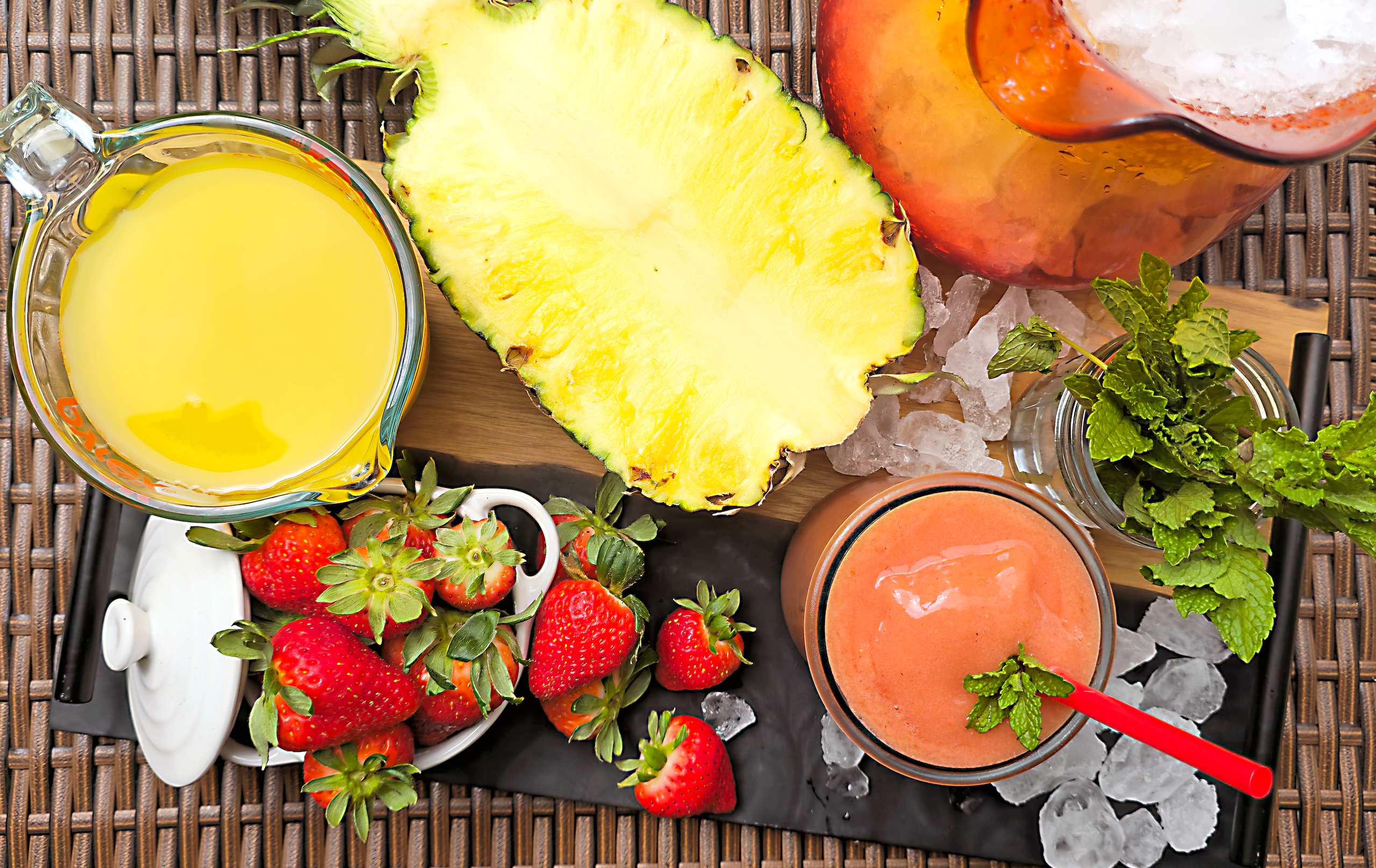 The strawberry fields smoothie begins with this overhead view of fresh strawberries in a white dish, a halved pineapple, a glass measuring cup filled with orange juice, a jar of fresh mint, and a peach-colored smoothie in a glass with a red straw and mint garnish, all arranged on a wooden board with scattered ice.