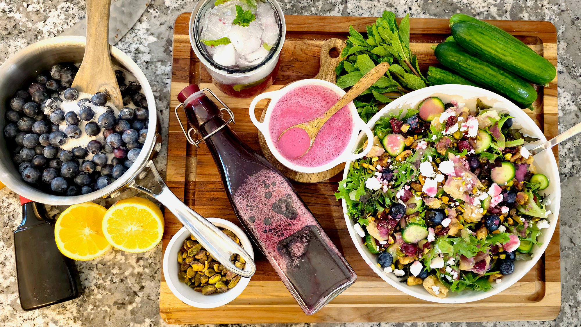 GRO-Blueberry Syrup.1 A wooden board displays fresh ingredients and a finished blueberry salad: a saucepan of blueberries being stirred with a wooden spoon, a bottle and small bowl of bright pink blueberry dressing, a glass of iced mint water, sliced lemons, pistachios, fresh mint, cucumbers, and a large bowl of mixed greens topped with blueberries, cucumbers, nuts, and crumbled cheese.