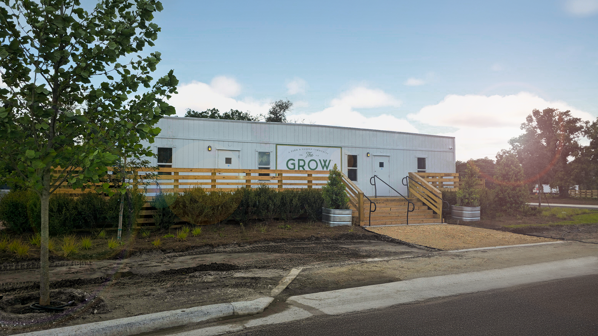Exterior view of welcome center where new home sales underway at The Grow — a white farmhouse-style building with wooden stairs, ramp access, landscaped greenery, and “The Grow” signage on the front under a bright blue sky.