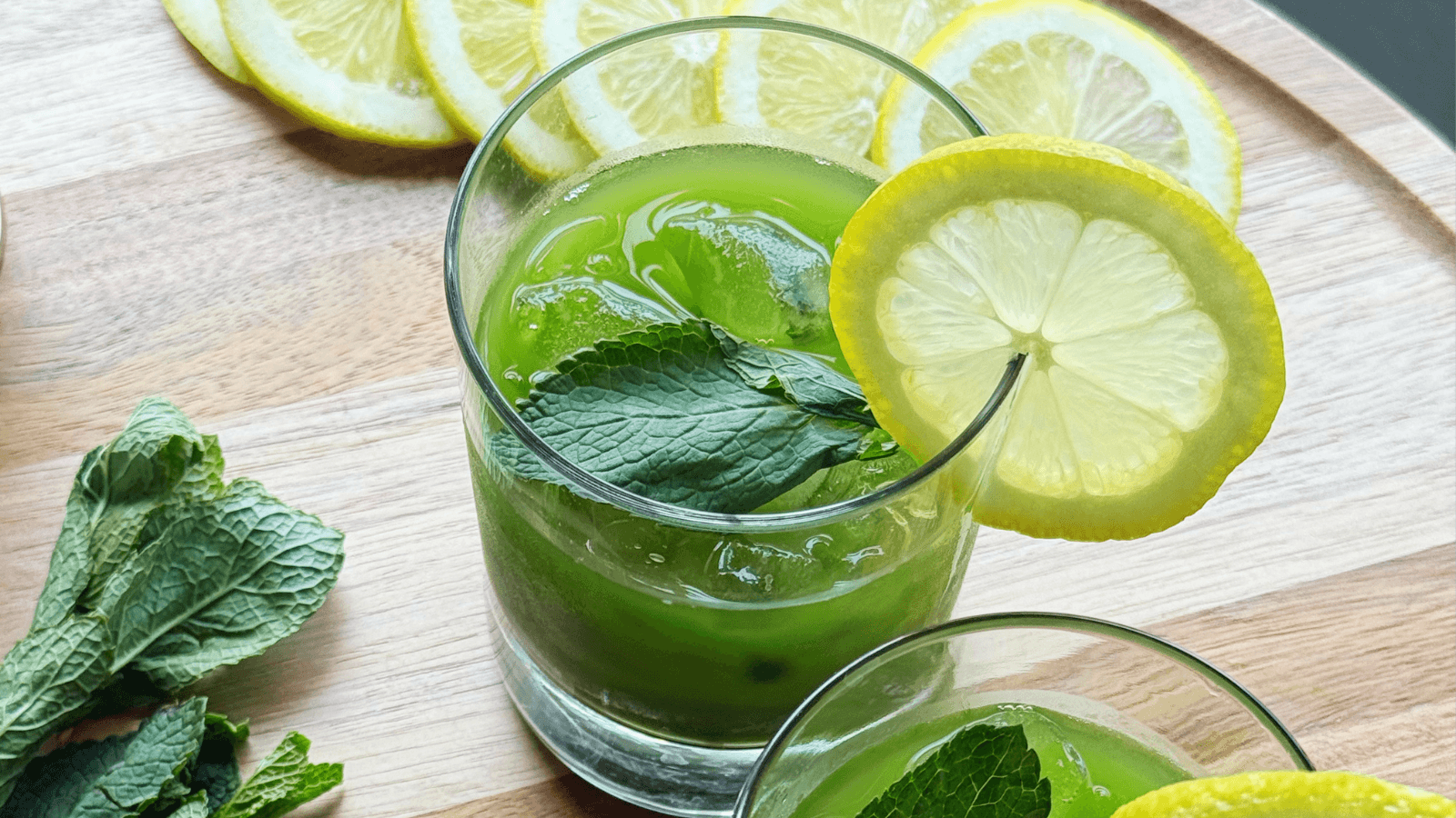 A refreshing green matcha cooler garnished with fresh mint and lemon slices, served over ice in a glass on a wooden cutting board. Fresh mint leaves and lemon slices are artfully arranged on the board.